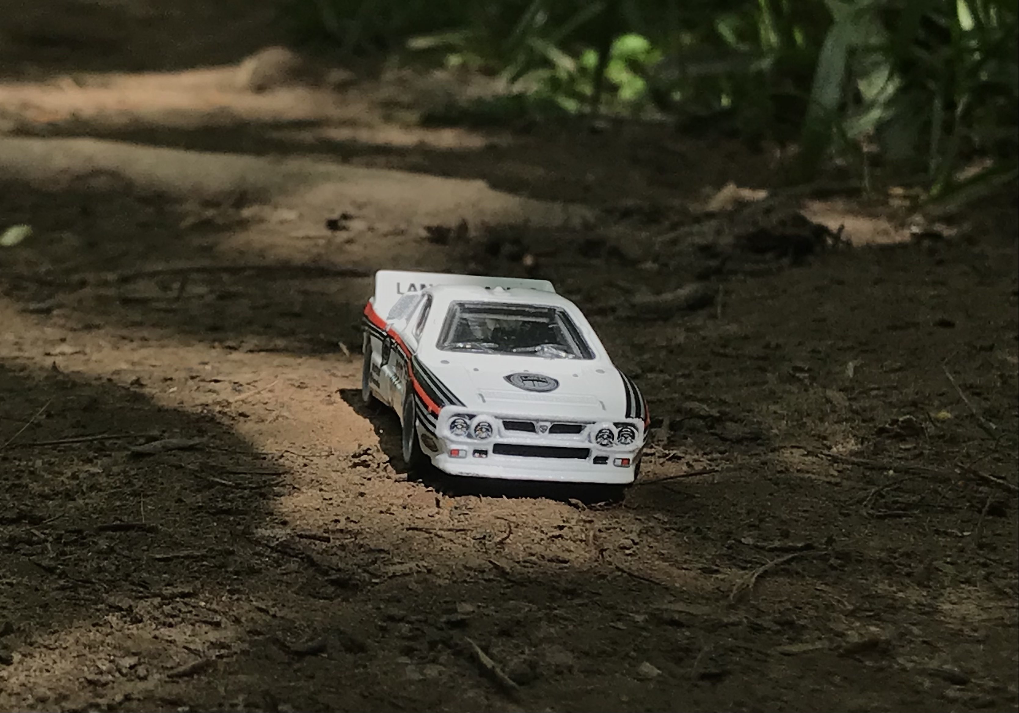 Hotwheels Grp B Rally Lancia 037 on a dirt path, with some grass in the background. The car is illuminated by sunlight, however is surrounded by shadows cast by leaves of trees overhead [World of Tanks - Main Menu 2]