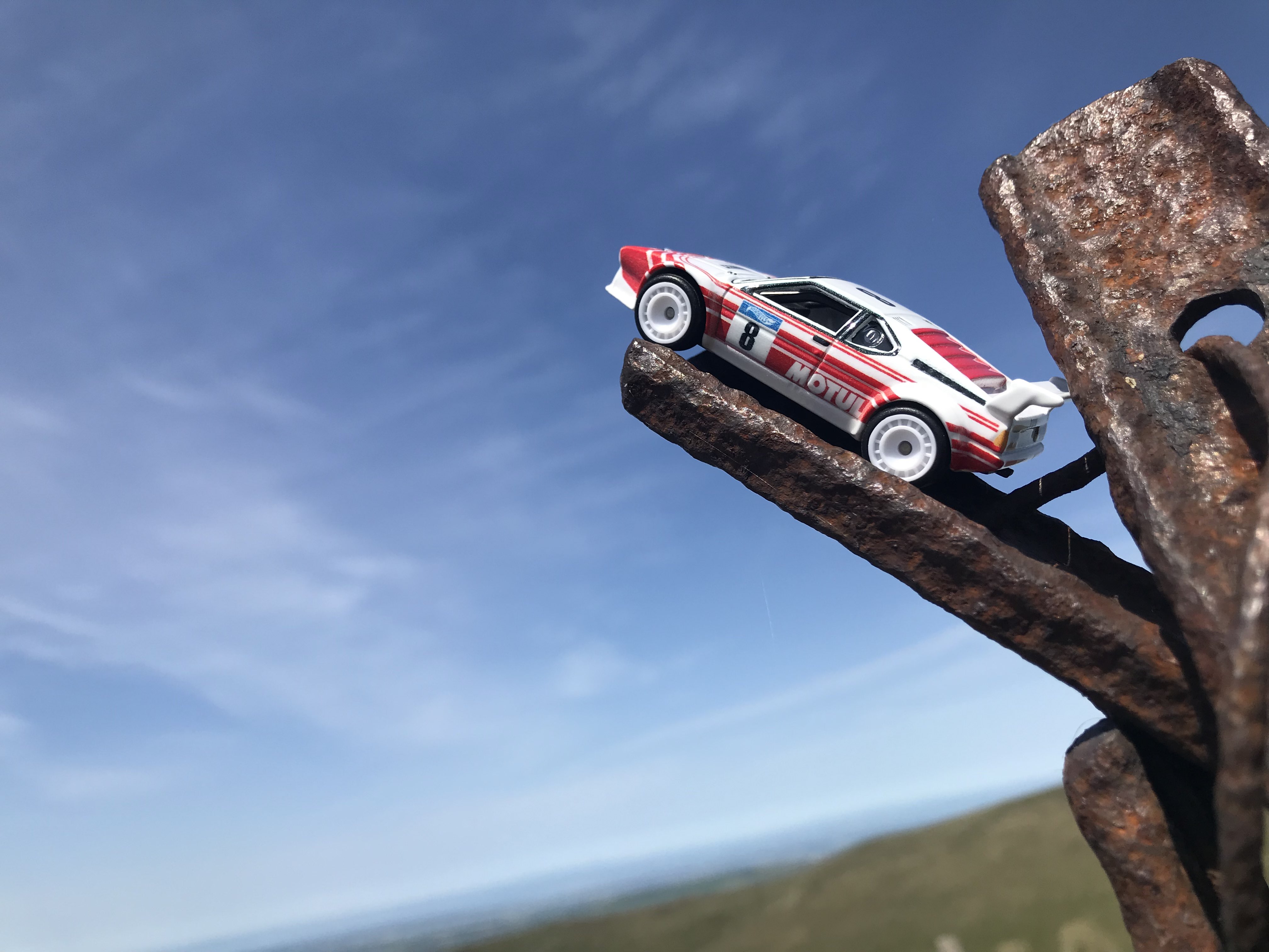 Hotwheels GrpB Rally BMW M1 on the end of a rusty metal bar, pointing into the sky. The horizon is at an angle, and most of the background is a clear sky [Noistcontrollers - Revolution is Here]