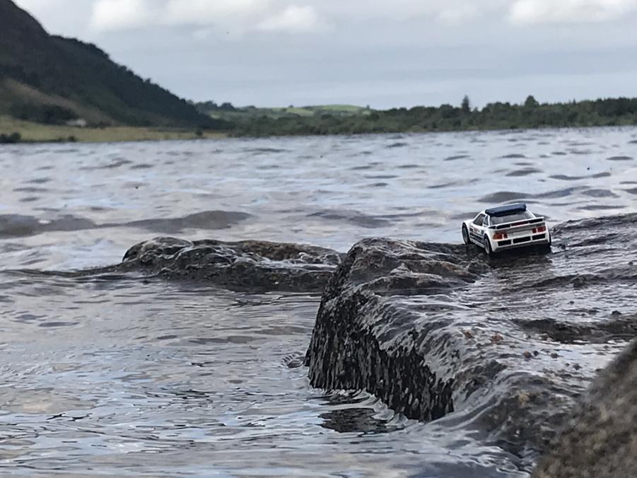Hotwheels Grp B Rally Ford RS200, on a rock surrounded by water. The water is part of a lake shown in the background. The other end of the lake is shown in the distance [Alphaville - Big in Japan]